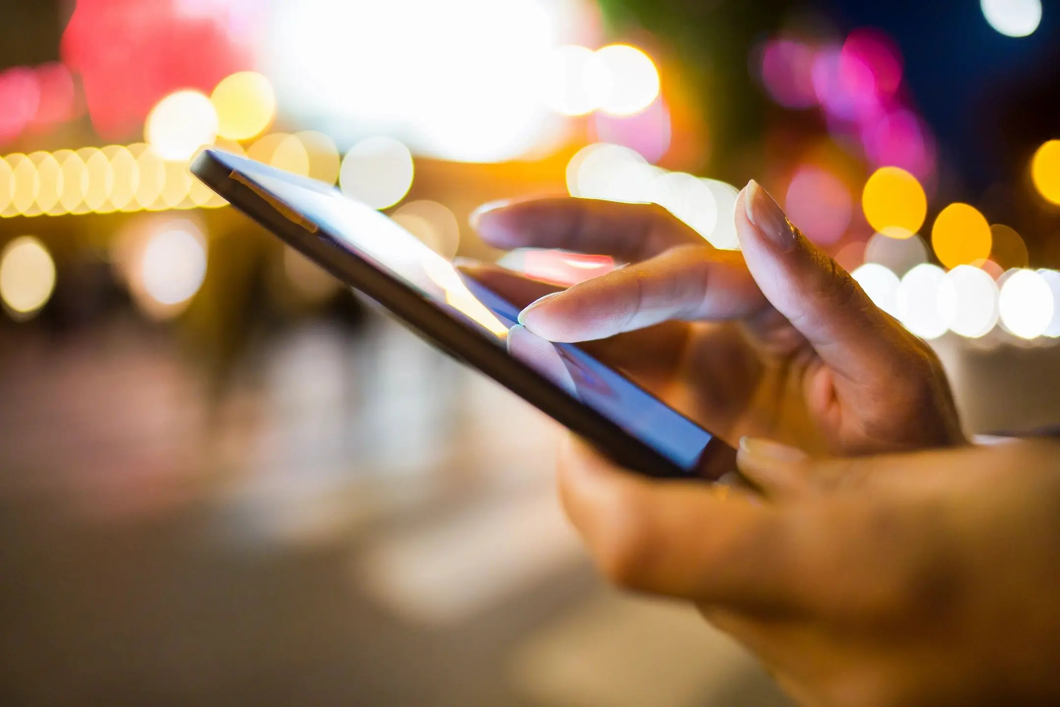 Person using a smartphone at night with blurred city lights in the background.
